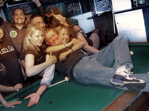 Happy pool team celebrates a great victory with their winning teammate being embraced with hugs and open arms while lying on the pool table.